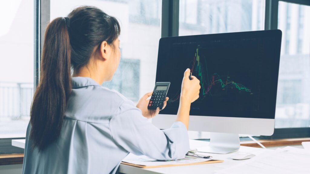 Woman sitting in front of computer with graphs using a calculator.