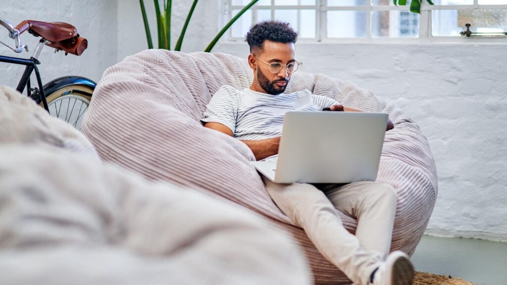 African American man sitting on beanbag working on a laptop.