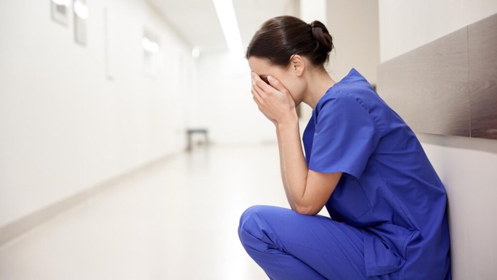 Nurse crouched by a wall in a hospital sad & burnt out because of declining mental health.