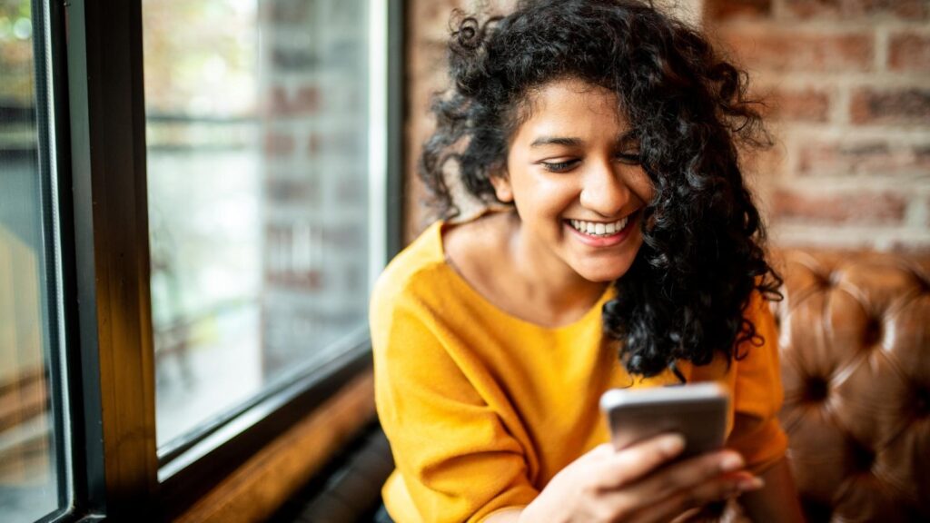 Woman at a cafe looking excited and smiling at her mobile phone.