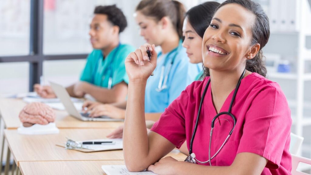 African American nurse in pink medical scrubs sitting in a classroom smiling at the camera.