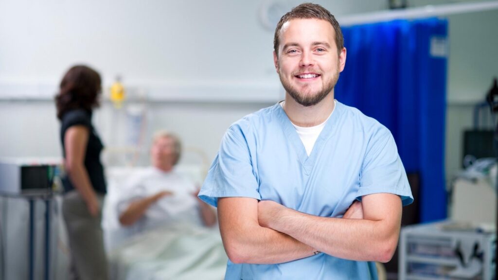 Male nurse in blue medical scrubs with a hospital background smiling with his arms crossed.