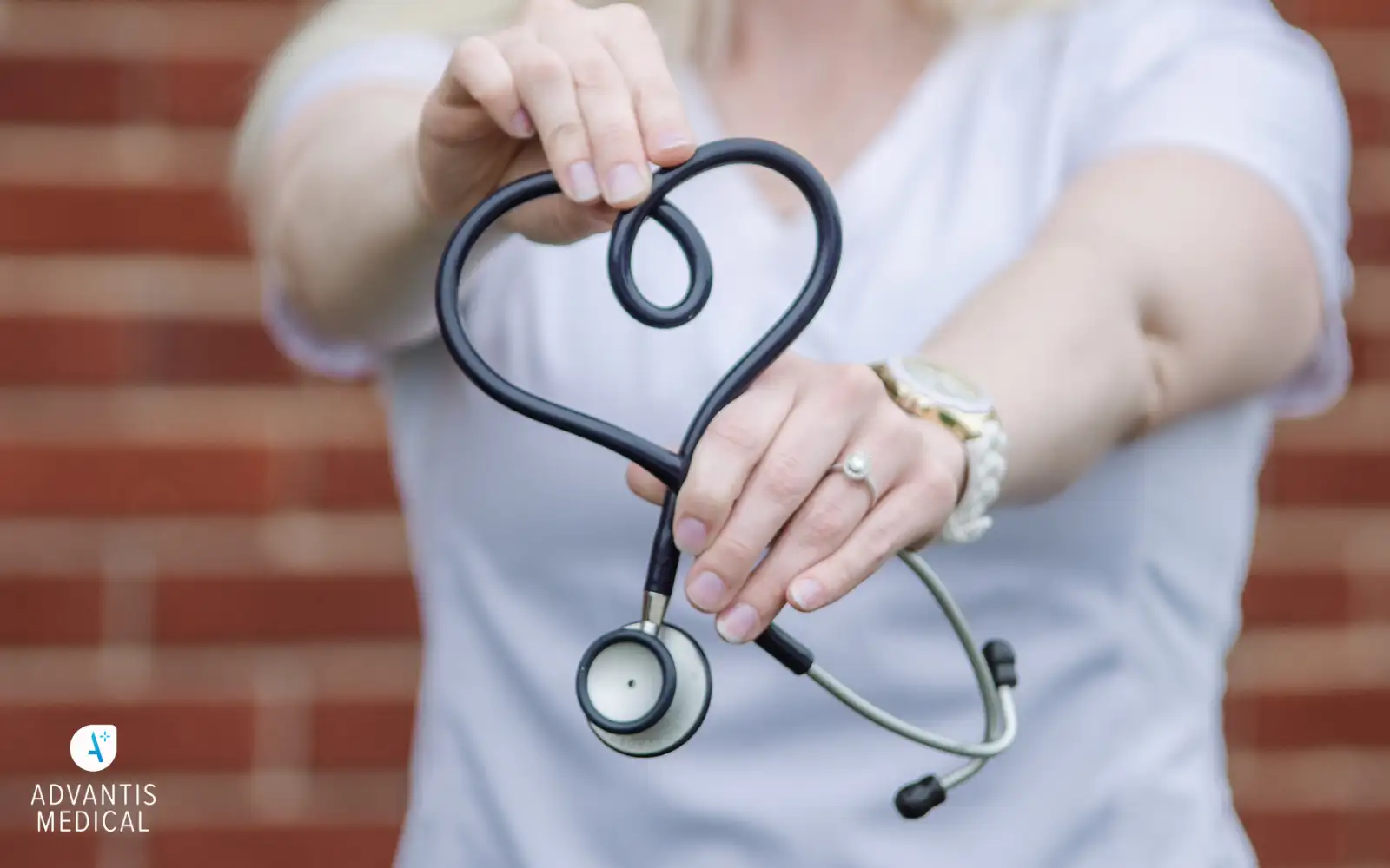 Woman wearing a white t shirt in front of red bricks holding a stethoscope in the shape of a heart.