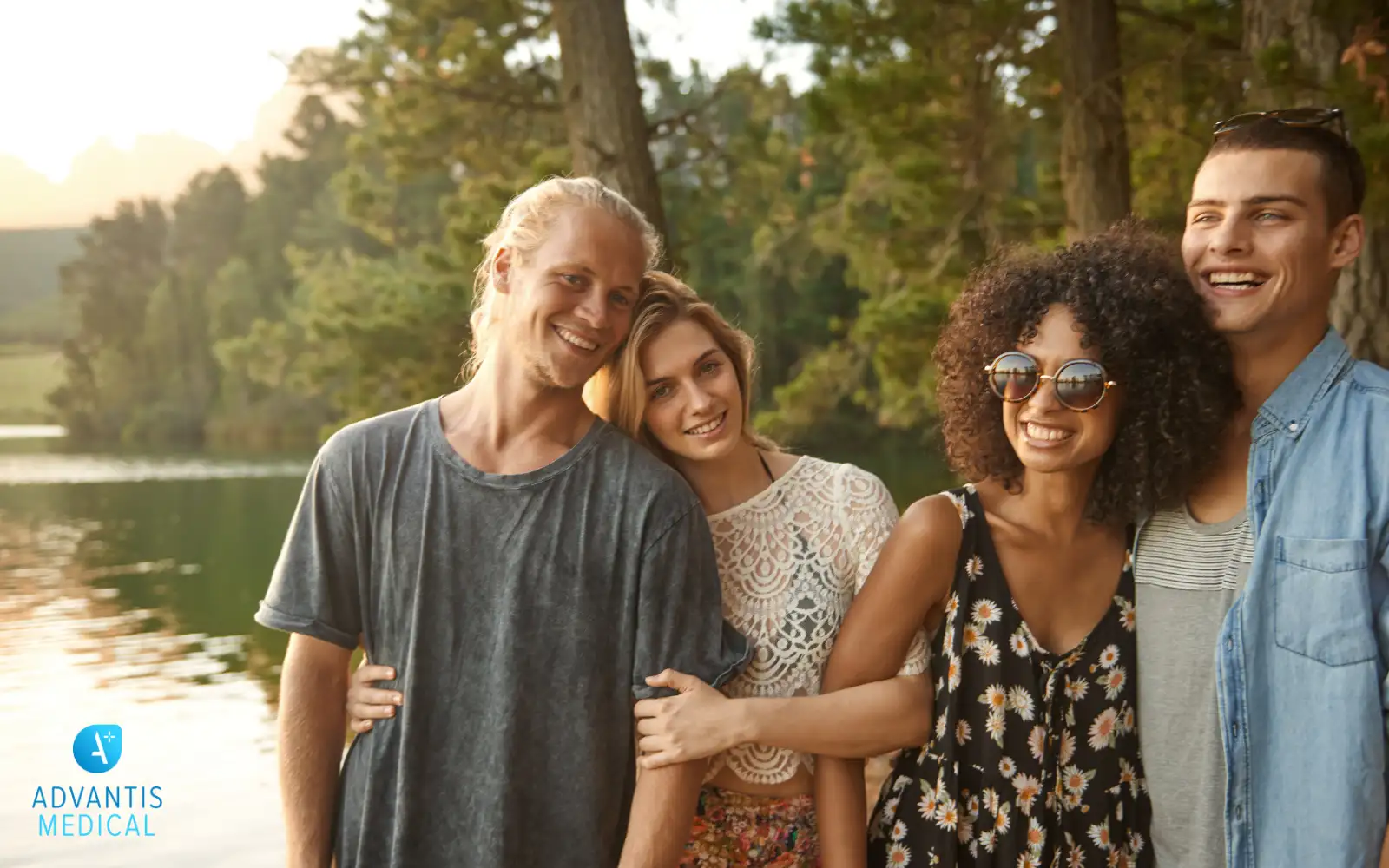 Group of diverse multi-cultural friends smiling and linking hands outdoors by a lake at sunset.