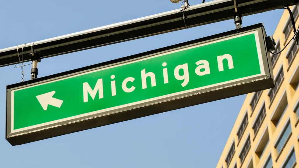Close up of green street sign with 'Michigan' written in white block text hanging with a sky and building in the background. Travel nurse housing in Michigan.