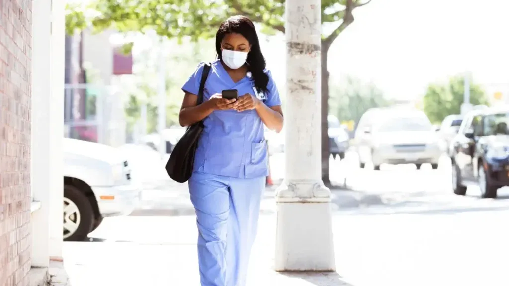 Female local travel nurse in blue medical scrubs wearing a mask on the phone while walking to work.